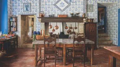 Французская кухня со старинным столом и стульями, терракотовой плиткой, стенами, выложенными синей плиткой, большим камином, медными кастрюлями, приставным столиком с фруктами и овощами, ступеньками направо наверх. Французская кухня со старинным столом и стульями, терракотовой плиткой, стенами, выложенными синей плиткой, большим камином, медными кастрюлями, приставным столиком с фруктами и овощами, ступеньками направо наверх.