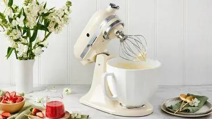 Кремовый миксер Kitchenaid Artisan с белой керамической чашей в окружении букета белых цветов, сока и набора тарелок. Кремовый миксер Kitchenaid Artisan с белой керамической чашей в окружении букета белых цветов, сока и набора тарелок.